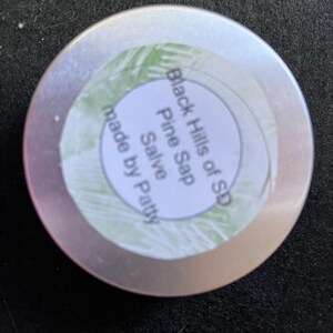 May include: A silver tin with a white label that reads "Black Hills of SD Pine Sap Salve made by Patty".