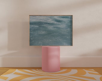 May include: A framed print of a blue ocean with waves, sitting on a pink cylindrical pedestal, on top of a yellow and white patterned rug.