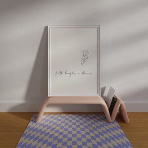 May include: A white framed print with a line drawing of a flower and the text "Earth laughs in flowers". The print is on a light pink shelf with a book on the shelf. The shelf is on top of a purple and white checkered rug.