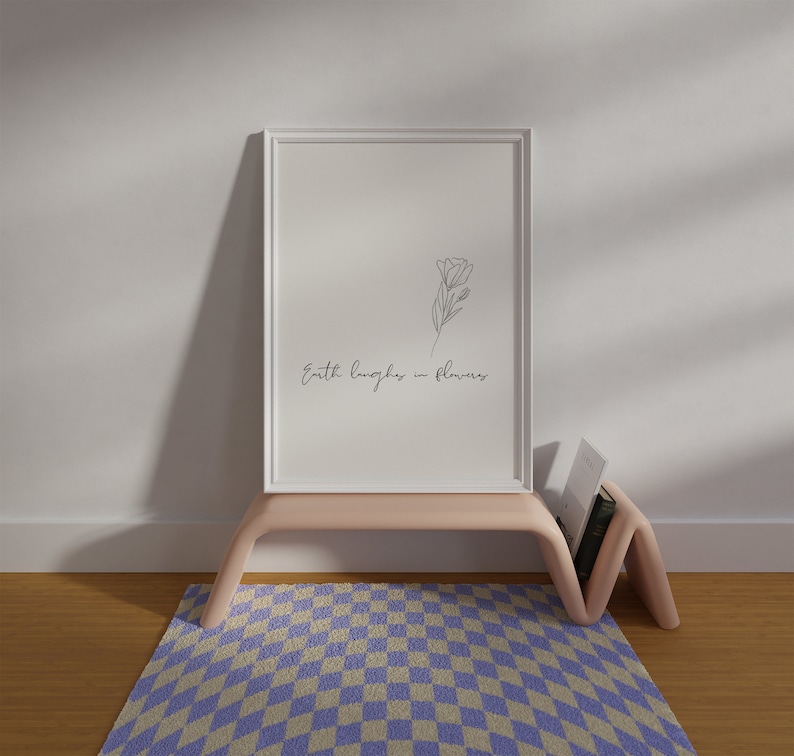 May include: A white framed print with a line drawing of a flower and the text "Earth laughs in flowers". The print is on a light pink shelf with a book on the shelf. The shelf is on top of a purple and white checkered rug.