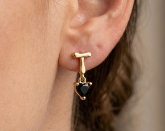 Könnte beinhalten: Ein goldener Ohrstecker mit einem großen Buchstaben T als Pfosten und einem baumelnden, herzförmigen schwarzen Edelsteinanhänger. Der Ohrring ist am Ohrläppchen befestigt und zeigt ein poliertes Metallfinish sowie einen facettierten dunklen Stein.
