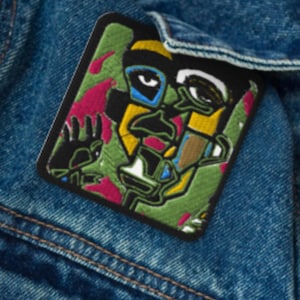 May include: A colorful embroidered patch with a geometric design featuring a face with large eyes, a nose, and a mouth. The patch is on a denim background.