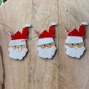 Könnte beinhalten: Drei Holzornamente in Form von Weihnachtsmannköpfen, jedes mit einem roten Hut, einem weißen Bart und schwarzen Augen. Die Ornamente hängen an Schnüren.