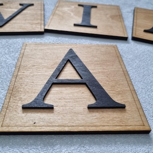 May include: Wooden letter tiles with a dark stain. The letter "A" is in focus, with other letters visible in the background. The letters are cut out of the wood and have a black border.