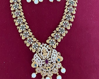 May include: Gold-toned necklace and matching earrings set. The necklace features a pendant with a central ruby and pearl drops. The earrings have a similar design with pearls. The set is adorned with white and colored gemstones.