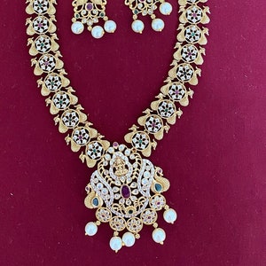 May include: Gold-toned necklace and matching earrings set. The necklace features a pendant with a central ruby and pearl drops. The earrings have a similar design with pearls. The set is adorned with white and colored gemstones.