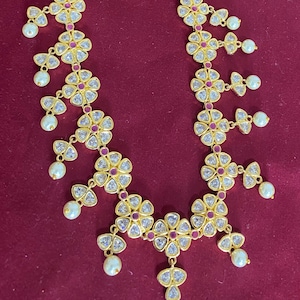 May include: A gold-toned necklace and matching earrings set. The necklace features three strands of small, deep red beads, with floral pendants adorned with clear stones and red accents. White pearls dangle from the pendants. The earrings have a similar floral design.