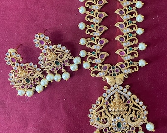 May include: Gold-toned jewelry set featuring a necklace and earrings. The necklace has a pendant with a deity figure and is adorned with red, green, and white gemstones, and faux pearls. The earrings mirror the necklace design.