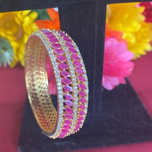 May include: A gold-toned bangle bracelet with a row of oval, magenta-colored stones and a row of clear, sparkling stones. The bracelet is displayed on a black stand against a backdrop of colorful flowers.