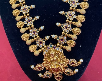 May include: Gold-toned necklace with intricate detailing. The necklace features a central pendant with a figure, surrounded by colorful gemstones and pearls. The design includes repeating floral and geometric patterns.