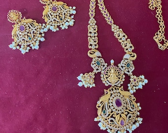 May include: A gold-toned jewelry set featuring a necklace and matching earrings. The necklace has a long chain with red and clear stone accents, and a large pendant with a central red stone, surrounded by clear stones, green stones, and white pearls. The earrings mirror the pendant design.