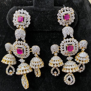 May include: A pair of gold-toned chandelier earrings. Each earring features a square, ruby-colored stone at the center of a square frame, surrounded by clear, sparkling stones. The design includes bell-shaped dangles and teardrop accents, all set against a black background.
