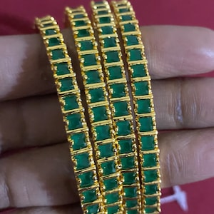 May include: Four gold-colored bangles with emerald green square-cut stones. The bangles are stacked together, showcasing the intricate design and the vibrant green color of the stones. The jewelry is displayed against a soft background.