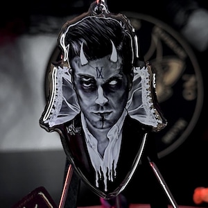 Peut inclure: Un porte-clés avec un portrait d'une personne avec des cornes de diable et un col blanc. Le porte-clés est attaché à un anneau en métal argenté. Une planche Ouija noire avec des lettres dorées est au premier plan. L'arrière-plan est sombre avec des éléments décoratifs.