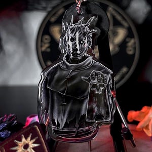 Puede incluir: Un llavero acrílico en blanco y negro con un personaje con rostro de calavera y una mano sosteniendo una calavera. El llavero está sujeto a un llavero plateado. El fondo incluye una tabla Ouija y flores secas.