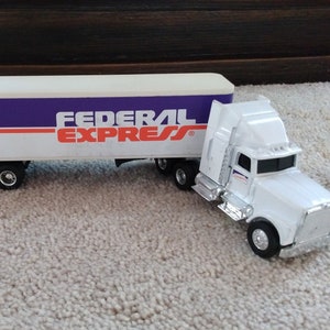 May include: A white toy semi-truck with a trailer. The trailer has a purple and orange "Federal Express" logo.