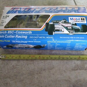 K&ouml;nnte beinhalten: Eine blaue und wei&szlig;e Schachtel f&uuml;r ein 1/24-Modell aus Metallguss eines March 85C-Cosworth IndyCar. Die Schachtel zeigt ein Foto des Autos mit dem Mobil 1 Racing-Logo und dem Text "March 85C-Cosworth Team Cotter Racing".