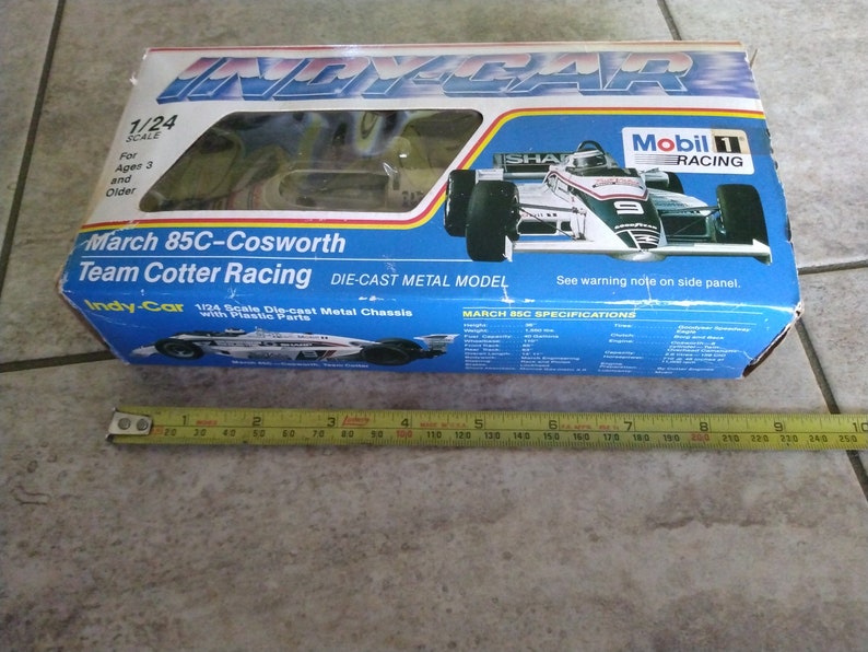 K&ouml;nnte beinhalten: Eine blaue und wei&szlig;e Schachtel f&uuml;r ein 1/24-Modell aus Metallguss eines March 85C-Cosworth IndyCar. Die Schachtel zeigt ein Foto des Autos mit dem Mobil 1 Racing-Logo und dem Text "March 85C-Cosworth Team Cotter Racing".