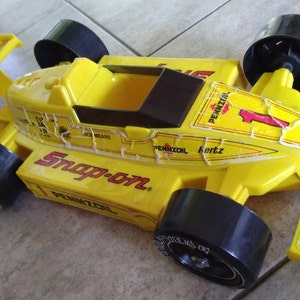 K&ouml;nnte beinhalten: Gelbes Spielzeug-Rennwagen mit schwarzen R&auml;dern und einem Snap-on-Logo. Das Auto hat Pennzoil-Branding und die Nummer 1 an der Seite.
