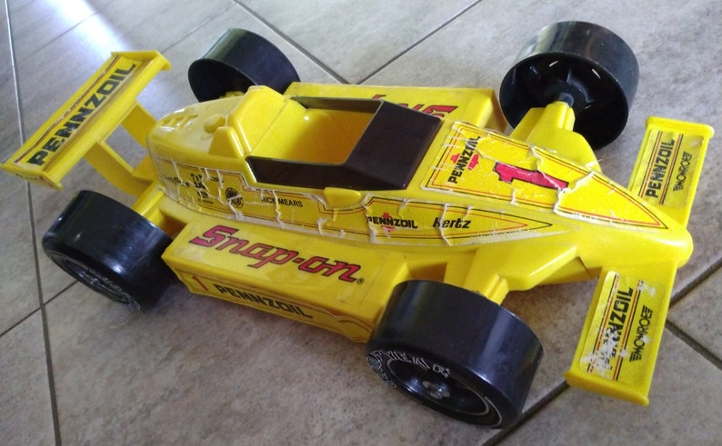 K&ouml;nnte beinhalten: Gelbes Spielzeug-Rennwagen mit schwarzen R&auml;dern und einem Snap-on-Logo. Das Auto hat Pennzoil-Branding und die Nummer 1 an der Seite.