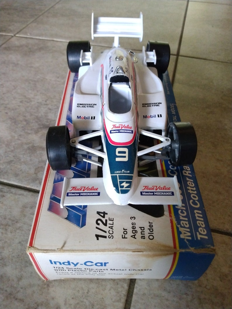 K&ouml;nnte beinhalten: Ein 1/24-Modell eines Indy-Rennwagens aus Druckguss mit wei&szlig;er Karosserie, blauen und roten Akzenten und den Worten "True Value Master Mechanic" an den Seiten. Das Auto befindet sich auf einem blauen und gelben Karton mit den Worten "Team Cotter Racing" und "March" an der Seite.