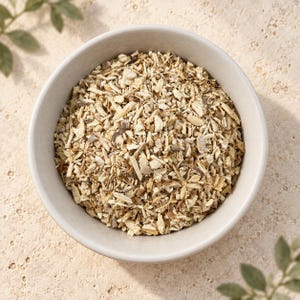 May include: A round, light-colored ceramic bowl filled with a heap of dried, chopped herbal root pieces. The contents consist of small, irregular, woody fragments in shades of beige, tan, and light brown, creating a textured, granular appearance.