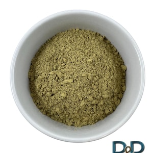 Peut inclure: Un bol en céramique blanc rempli d'une poudre finement moulue, vert olive. La poudre a un aspect légèrement texturé. Le bol est centré sur un fond blanc uni. L'image est une vue de dessus.