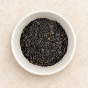 May include: A white ceramic bowl filled with a coarse, dark brown and black granular substance. The mixture contains small, irregular particles, dark soil-like fragments, and tiny bits of organic matter, creating a textured, earthy appearance.