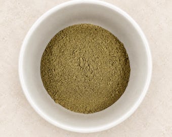 Puede incluir: Un cuenco de cerámica blanca lleno de un polvo fino de color verde oliva terroso. El cuenco es redondo con bordes lisos y limpios, y el contenido está distribuido uniformemente, mostrando una textura mate y consistente en la superficie.