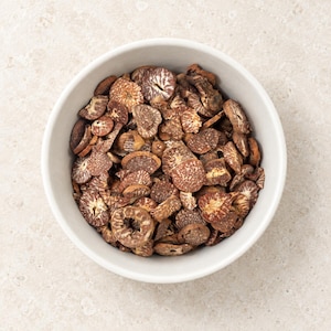 May include: A white bowl filled with numerous small, dried, sliced betel nuts. The slices are circular and irregular in shape, displaying a textured, mottled brown and tan surface with visible fibrous patterns throughout each individual piece.
