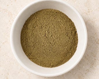 Puede incluir: Un cuenco redondo de cerámica blanca lleno de un polvo fino de color verde oliva terroso. El polvo tiene una textura mate y uniforme que cubre toda la base interior del cuenco, creando una superficie circular plana de material molido.
