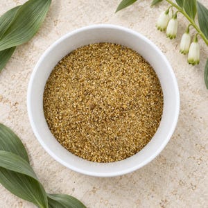 May include: A white ceramic bowl filled with a coarse, golden-brown granular substance. The texture appears dry and finely ground, featuring a mix of light and dark brown particles throughout the mixture, contained within a simple, round vessel.