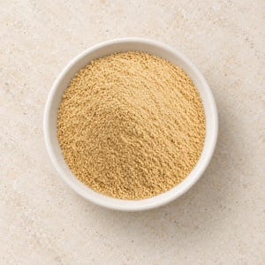 May include: A small, round, white ceramic bowl filled with a mound of fine, light tan, granular powder. The texture of the powder appears uniform and dry, with a slightly coarse consistency, neatly contained within the circular rim of the bowl.