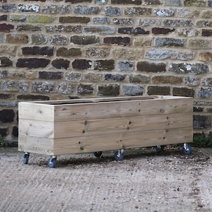May include: A wooden planter box on wheels. The planter is made of natural wood and has a rustic look. It is sitting in front of a brick wall.