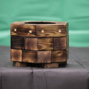 May include: A square wooden planter box with a dark brown finish. The planter has a rustic look and is made from multiple pieces of wood.