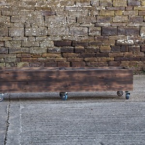 May include: A dark brown wooden planter box on wheels. The planter is made of reclaimed wood and has a rustic finish. The wheels are black metal with blue rubber tyres.
