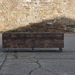 May include: A dark brown wooden planter box on small caster wheels. The rectangular box is made of stained wood and is designed for outdoor use. The background features a weathered brick wall and a concrete surface.