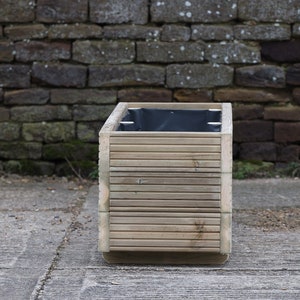 May include: A wooden planter box with a black liner. The planter is made of natural wood and has a slatted design. The planter is sitting on a concrete surface.
