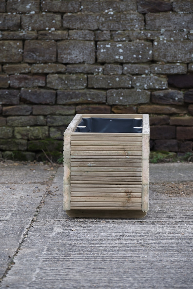 May include: A wooden planter box with a black liner. The planter is made of natural wood and has a slatted design. The planter is sitting on a concrete surface.