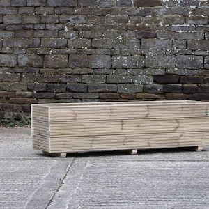 May include: A long, rectangular wooden planter box with a slatted design. The planter is made of natural wood and has a light brown colour. It is sitting on a concrete surface in front of a brick wall.