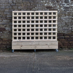 May include: A wooden planter box with a trellis on the back. The planter is on wheels and has a natural wood finish.