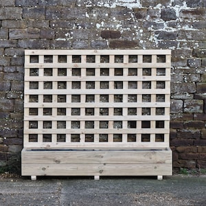 May include: A wooden trellis planter box. The planter box is made of light-coloured wood and features a lattice design. The box is rectangular and sits on small feet. The background is a weathered brick wall.