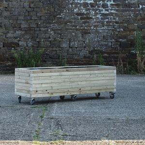 May include: A wooden planter box on wheels. The planter is made of natural wood and has a dark brown liner. The planter is sitting on a grey concrete surface.