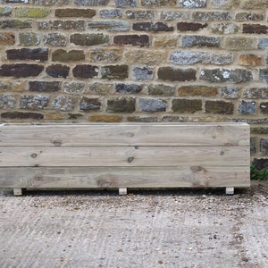 May include: A long, rectangular wooden planter box with three horizontal planks. The planter is sitting on a grey concrete surface in front of a stone wall.