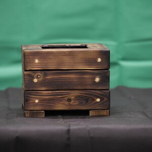 May include: A dark brown wooden planter box with three tiers. The planter has a square shape and is made of wood planks. The planter has brass accents on the corners.