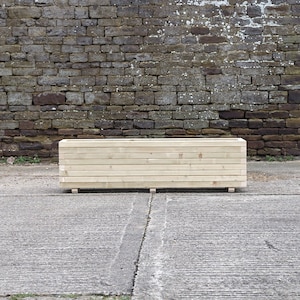 May include: A long, rectangular wooden planter box with a natural finish. The planter is made of horizontal wooden planks and sits on a concrete surface in front of a brick wall.