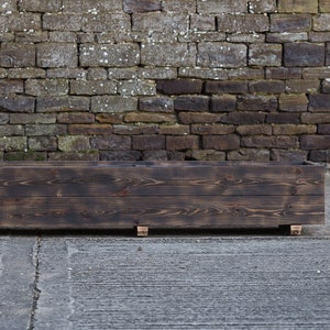 May include: A dark brown wooden planter box with a rectangular shape. The planter is sitting on a grey concrete surface in front of a brick wall.