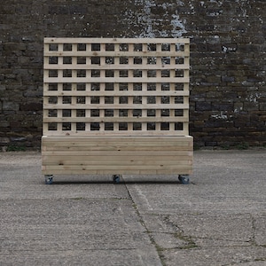 May include: A wooden planter box with a trellis on the back. The planter is on wheels and has a natural wood finish.