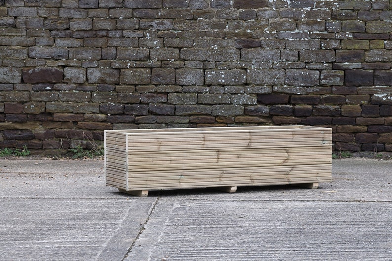 May include: A rectangular wooden planter box with a natural finish. The planter is made of horizontal slats of wood and has a simple design.