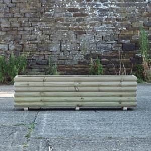 May include: A long, rectangular wooden planter with a rounded top and a natural wood finish. The planter is sitting on a concrete surface in front of a brick wall.